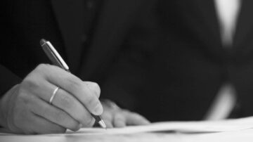رجل يرتدي بدلة يكتب بقلم على ورقة، يظهر التركيز في يده التي تحمل القلم بينما يكون جزء من جسمه في الخلفية.