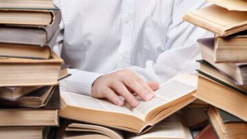 رجل يقرأ كتابًا مفتوحًا محاطًا بأكوام من الكتب المكدسة حوله. تركز الصورة على اليد التي تلمس صفحات الكتاب، مما يعكس حب القراءة والمعرفة.