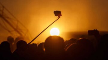 صورة تظهر مجموعة من الأشخاص في ظلال silhouetted يحملون عصا سيلفي، مع غروب الشمس في الخلفية. الأضواء البرتقالية والصفراء تخلق جوًا دافئًا ومثيرًا.