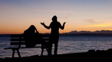 شخصان silhouetted على ضفاف المياه خلال غروب الشمس، أحدهما يجلس على مقعد والآخر واقف يعبر عن مشاعره. الأفق يظهر الجبال تحت ضوء الغروب الدافئ.