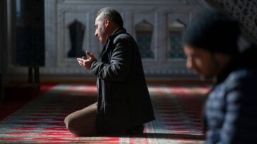 رجل يؤدي الصلاة في مسجد، جاثيًا على ركبتيه في وضع خشوع، بينما يظهر رجل آخر في الخلفية. الإضاءة الطبيعية تضيف جوًا هادئًا للمشهد.