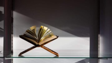 كتاب مفتوح على حامل خشبي، مع ظلال خفيفة تسلط الضوء على الصفحات. يتميز التصميم البسيط بواجهة زجاجية تظهر الكتاب بشكل أنيق.