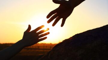 يد تمتد نحو الشمس أثناء غروبها، مع وجود يد أخرى تقترب منها، تعكس لحظة تواصل وأمل.