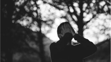 رجل يقف في الطبيعة، يضع يديه على وجهه في تعبير عن الانزعاج أو الحزن، مع خلفية ضبابية لأشجار مظللة. الصورة بالأبيض والأسود تعكس شعوراً عميقاً من العزلة.