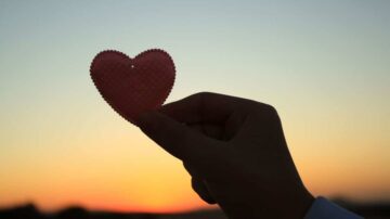 يد تحمل قلبًا ورديًا أمام غروب الشمس، مع ألوان دافئة في السماء. يرمز المشهد إلى الحب والأمل.
