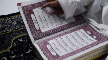 كتاب مقدس مفتوح مع يد تشير إلى النص، يظهر آيات مكتوبة بخط عربي جميل. الخلفية تحتوي على سجادة صلاة تقليدية، مما يبرز الأجواء الروحية.