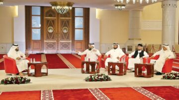 خمس شخصيات في مناقشة حوارية داخل قاعة مزينة بسجاد أحمر، يجلسون حول طاولات خشبية صغيرة. يظهر في الخلفية أبواب مزخرفة وأضواء متلألئة. الجمع يتكون من رجال يرتدون الزي التقليدي العربي.