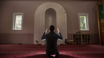 رجل يصلي في مسجد، يجلس على السجادة الحمراء مع رفع يديه للدعاء أمام المحراب، مع نوافذ مضيئة في الخلفية.