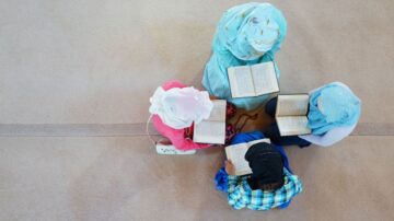 أربع نساء يرتدين أزياء تقليدية، يجلسن على الأرض في حلقة، يقرأن كتبًا مفتوحة. تظهر الألوان الزاهية لأغطية الرأس والملابس، مما يخلق جوًا من الهدوء والتركيز على التعليم والقراءة.