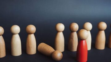 مجموعة من تماثيل خشبية صغيرة، حيث يظهر تمثال أحمر يقف بين تماثيل خشبية طبيعية، بينما يوجد تمثال خشبي مسقط على الأرض. الخلفية داكنة، مما يبرز الألوان. يمثل المشهد فكرة الفردية والتميّز.