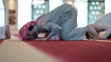 رجل يؤدى الصلاة في مسجد، مع التركيز على السجود على سجادة حمراء. يظهر في الخلفية مصلون آخرون.