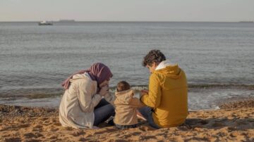 عائلة تتناول الطعام معًا على شاطئ البحر خلال غروب الشمس. يظهر الوالدان وطفلهما جالسين على الرمال، يستمتعون بلحظات هادئة مع المنظر الطبيعي للمحيط في الخلفية.