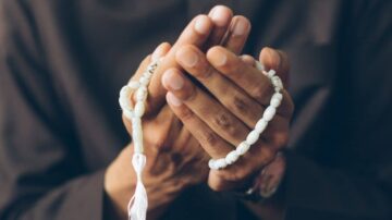 رجل يحمل سبحة بيضاء في يديه، مع التركيز على الأيدي المتشابكة. تعبر الصورة عن العبادة والتأمل الروحي.