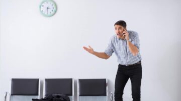 رجل يتحدث في الهاتف بقلق في غرفة انتظار، مع ساعة على الحائط تظهر الوقت. يظهر تعبيره على وجهه الإحباط، ويقف بجانب مقاعد فارغة.