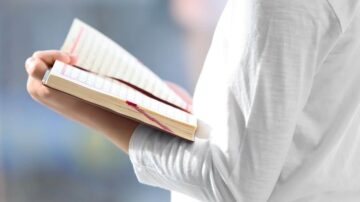 امرأة تحمل دفتر ملاحظات مفتوح، تظهر يدها وهي تمسك بالدفتر. يركز الضوء على الصفحات المكتوبة، مما يبرز محتوى الكتابة.