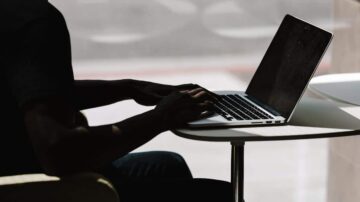 رجل يستخدم جهاز كمبيوتر محمول على طاولة صغيرة، مع التركيز على يديه أثناء الكتابة. الضوء الطبيعي يدخل من النافذة، مما يخلق ظلًا على الشاشة.