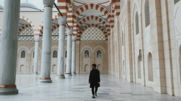 شخص يمشي في قاعة واسعة لمسجد، مع أعمدة رخامية وسقف مزخرف بقوسات حمراء وبيضاء. تعكس الأرضية اللامعة الضوء، مما يخلق جوًا هادئًا وجميلًا.