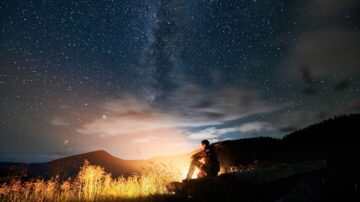 رجل يجلس بجانب نار مخيم تحت سماء مليئة بالنجوم، مع وجود درب التبانة واضحًا في الخلفية، وسط طبيعة جبلية.