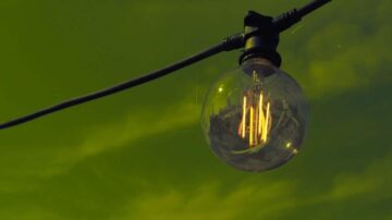 مصباح زجاجي معلق على سلك كهربائي، يظهر في الخلفية سماء بلون أخضر. يضيء المصباح بخيوط مضيئة، مما يخلق جوًا دافئًا.