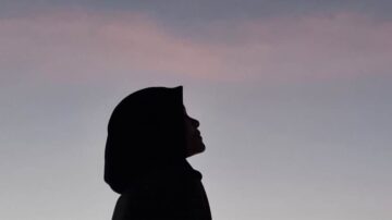 صورة ظلية لشخص يرتدي غطاء رأس، يتجه نظره نحو السماء مع ألوان غروب الشمس في الخلفية. تعكس الصورة لحظة تأمل وهدوء.