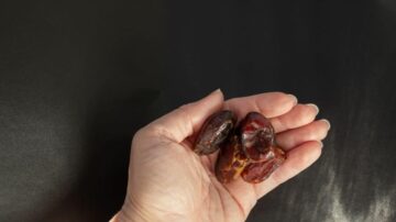 يد تحمل ثلاث حبات من التمر البني الداكن على خلفية سوداء، مع تركيز الضوء على التمر لإبراز تفاصيله وقوامه.