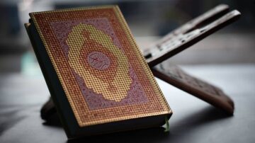 كتاب مزخرف بتصميم دقيق، موضوع على حامل خشبي، مع خلفية ضبابية، يعكس جمال الثقافة الإسلامية.