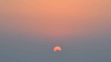 غروب شمس هادئ فوق الأفق، يظهر جزء من الشمس بلون برتقالي دافئ في سماء مائلة إلى اللون الوردي، مع ضباب خفيف يضيف لمسة من السلمية للمشهد.