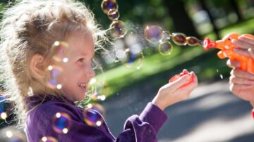 طفلة تلعب في الهواء الطلق وتبتسم بينما تُطلق فقاعات من لعبة فقاعات ملونة، مع أشعة الشمس تسطع حولها.
