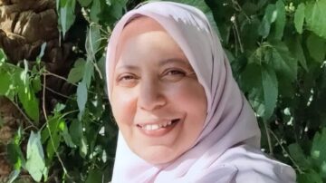 امرأة مبتسمة ترتدي حجابًا ورديًا، تقف أمام خلفية من أوراق خضراء وأشجار. تعكس الصورة شعورًا بالهدوء والسعادة.