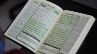 Open Quran displaying Arabic text on its pages, with intricate calligraphy and green borders. The book is positioned on a flat surface, highlighting its detailed layout.