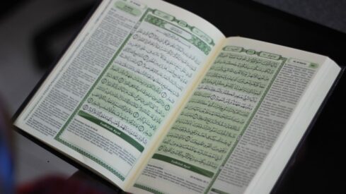 Open Quran displaying Arabic text on its pages, with intricate calligraphy and green borders. The book is positioned on a flat surface, highlighting its detailed layout.