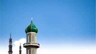 A green and gold dome of a mosque tower against a clear blue sky, featuring intricate architectural details. Several minarets are visible in the background, showcasing traditional Islamic design.