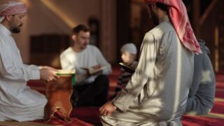 Men and children engaged in a Quran study session in a mosque, sitting on a red carpet. One man reads from a Quran while another observes. Soft lighting illuminates the scene, highlighting their focus and spirituality.