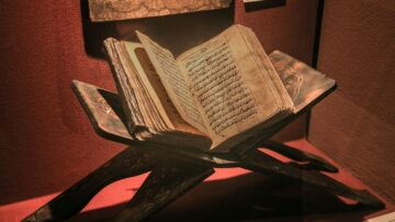 كتاب قديم مفتوح على حامل خشبي، يعرض نصوصاً مكتوبة بخط عربي تقليدي، محاط بجدران حمراء.