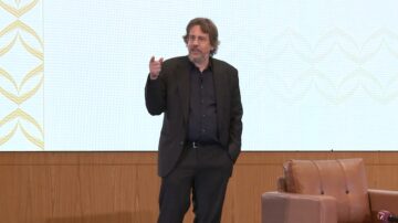 رجل يرتدي بدلة سوداء يقف على منصة ويتحدث إلى الجمهور. خلفه شاشة عرض كبيرة تعرض نمطًا هندسيًا. الأثاث من الجلد البني يتواجد بجانبه.