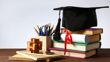 قبعة تخرج سوداء موضوعة على مجموعة من الكتب الملونة، مع أسطوانة ورقية مربوطة بشريط أحمر، وحاوية تحتوي على أقلام وأدوات مكتبية، بالإضافة إلى لعبة تركيب خشبية، ومفكرة ومسطرة، على سطح خشبي.