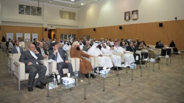 حضور كبير في قاعة مؤتمرات مع رجال أعمال ومشاركين في حدث. يتواجد رجال في بدلات تقليدية وحديثة، يجلسون على مقاعد مريحة. خلفية المكان تحتوي على شاشات عرض وديكور عصري.