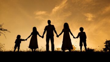 عائلة تتكون من خمسة أفراد، بما في ذلك أطفال، يقفون معًا ممسكين بأيدي بعضهم البعض في صورة ظلية ضد غروب الشمس. السماء تتلون بألوان دافئة، مما يخلق جوًا من الحب والترابط.