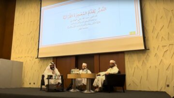 جلسة نقاشية حول تأثير القرآن الكريم، تضم ثلاثة متحدثين، مع عرض تقديمي خلفهم. تظهر في الصورة شاشتان كبيرتان وديكور داخلي حديث، مع تركيز على موضوع الدراسات الإسلامية.