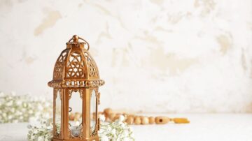 فانوس خشبي مزخرف بجانب زهور صغيرة وخرز خشبي على سطح أبيض مع خلفية دافئة. يبرز التصميم الأنيق والديكور البسيط، مما يخلق جوًا مريحًا.