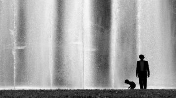 رجل وطفل يقفان أمام نافورة مياه، مع انبعاث المياه بشكل متساقط في الخلفية. الصورة باللونين الأبيض والأسود، تبرز الظلال والتباين بين الشخصين والمياه المتدفقة.