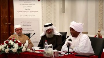 رجل يرتدي عمامة بيضاء يتحدث أمام ميكروفون في حدث رسمي، مع وجود رجلين آخرين بجانبه يرتديان ملابس تقليدية. خلفهم لافتات تتعلق بالثقافة العربية. الطاولة مزينة بالورد الأحمر وزجاجة ماء.