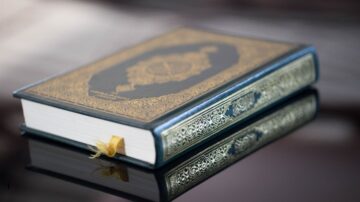 كتاب ذو غلاف مزخرف باللون الذهبي والأسود، يظهر على سطح لامع. يتضمن تصميمه آيات قرآنية، مما يبرز جمالياته الدينية والثقافية.