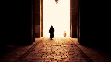 امرأتان تسيران عبر مدخل مظلم، مع إضاءة ساطعة في النهاية. الأرضية مبلطة بالحجارة، ويظهر ضوء خافت من الخارج. تعتبر الصورة تجسيدًا للتباين بين الظلام والنور، مما يرمز إلى الأمل والانتقال.