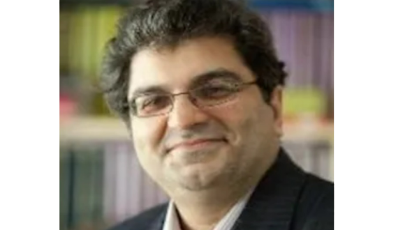 رجل في بدلة رسمية يبتسم أمام خلفية تحتوي على رفوف مملوءة بالكتب. الصورة تُظهر تفاصيل وجهه مع نظارات وشعر داكن ومجعد.