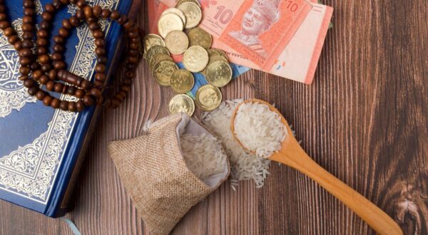 صورة تظهر كيس من الأرز مع ملعقة خشبية، إلى جانب أوراق نقدية ومجموعة من العملات المعدنية. خلفية الصورة خشبية، مع وجود مسبحة في الزاوية. تعكس الصورة مفاهيم الكرم والتبرع.