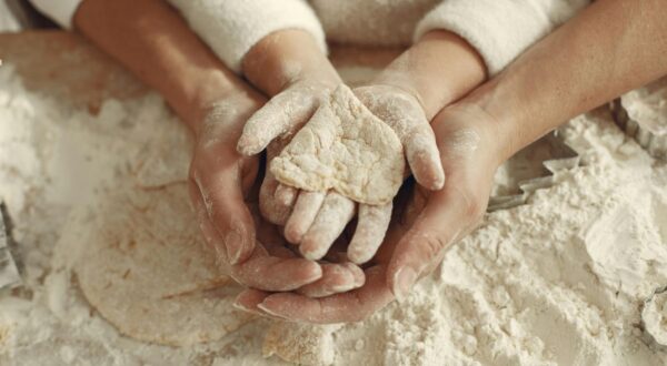 أيدٍ مغطاة بالدقيق تتداخل معًا، حيث تحمل يد صغيرة قطعة من العجين، تعبر عن اللحظات العائلية في المطبخ.