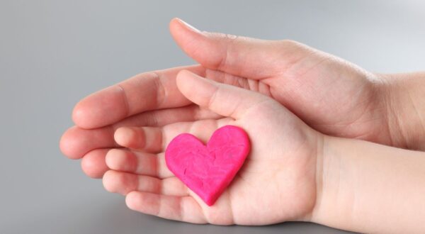 يدين تتصافحان مع قلب وردي مصنوع من الصلصال، توضح مشاعر الحب والمودة. خلفية رمادية.