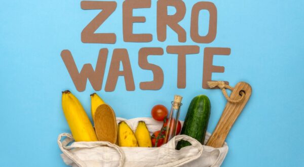 حقيبة قماشية تحتوي على فواكه وخضروات مثل الموز والخيار، مع كلمة "صفر نفايات" مكتوبة بأحرف كبيرة فوقها، على خلفية زرقاء.