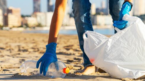 شخص يرتدي قفازات زرقاء يجمع النفايات من الشاطئ، ممسكًا بزجاجة بلاستيكية. يظهر في الخلفية مشهد للمدينة مع مبانٍ عالية.